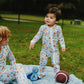 A toddler stands on a light blue quilted blanket in a park wearing a white long-sleeve footless pajama set with a colorful cartoon pattern. Another child is seated on the blanket, with a blue octopus toy and a football nearby.
