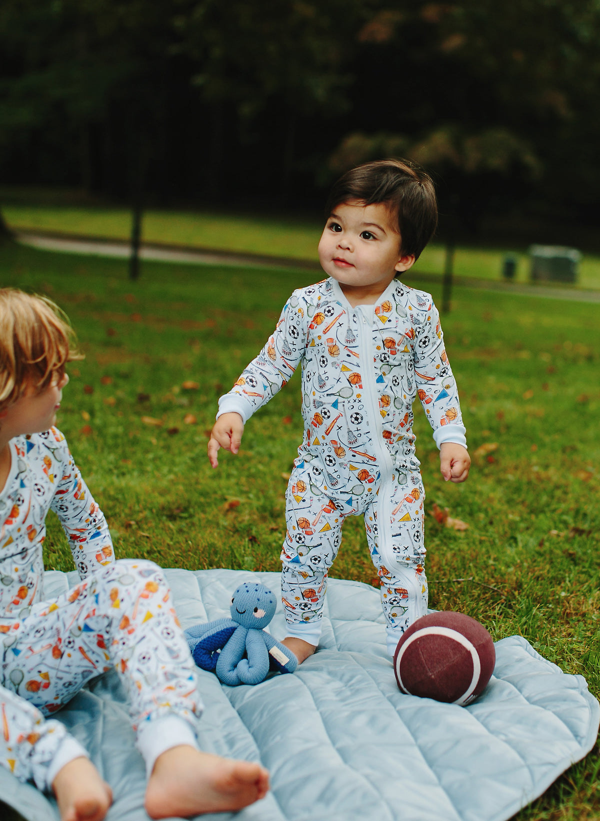 A toddler stands on a light blue quilted blanket in a park wearing a white long-sleeve footless pajama set with a colorful cartoon pattern. Another child is seated on the blanket, with a blue octopus toy and a football nearby.