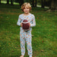A young child stands barefoot on a grassy lawn holding a football, wearing a long-sleeve pajama set with a colorful cartoon pattern on a light blue base.