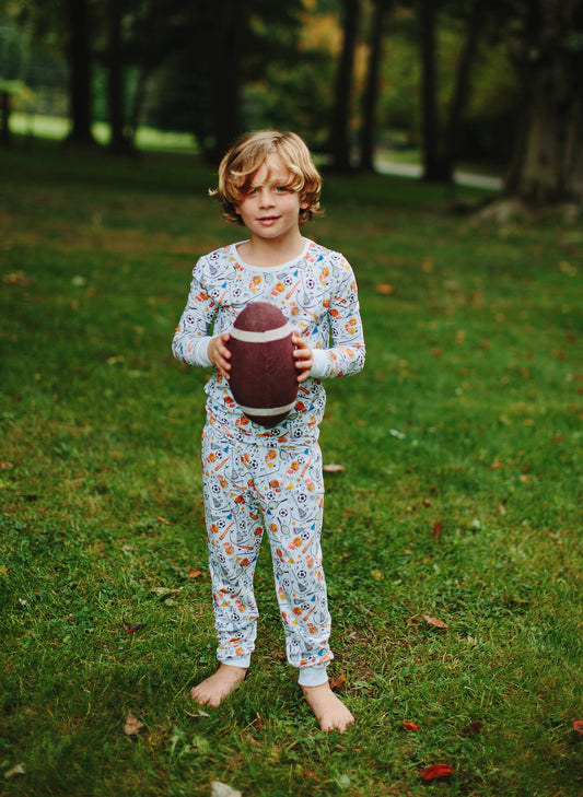 A young child stands barefoot on a grassy lawn holding a football, wearing a long-sleeve pajama set with a colorful cartoon pattern on a light blue base.