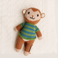A handmade knitted monkey toy with brown fur-like yarn, a striped green and blue sweater, and cream-colored feet and ears, resting on a textured white knit blanket.