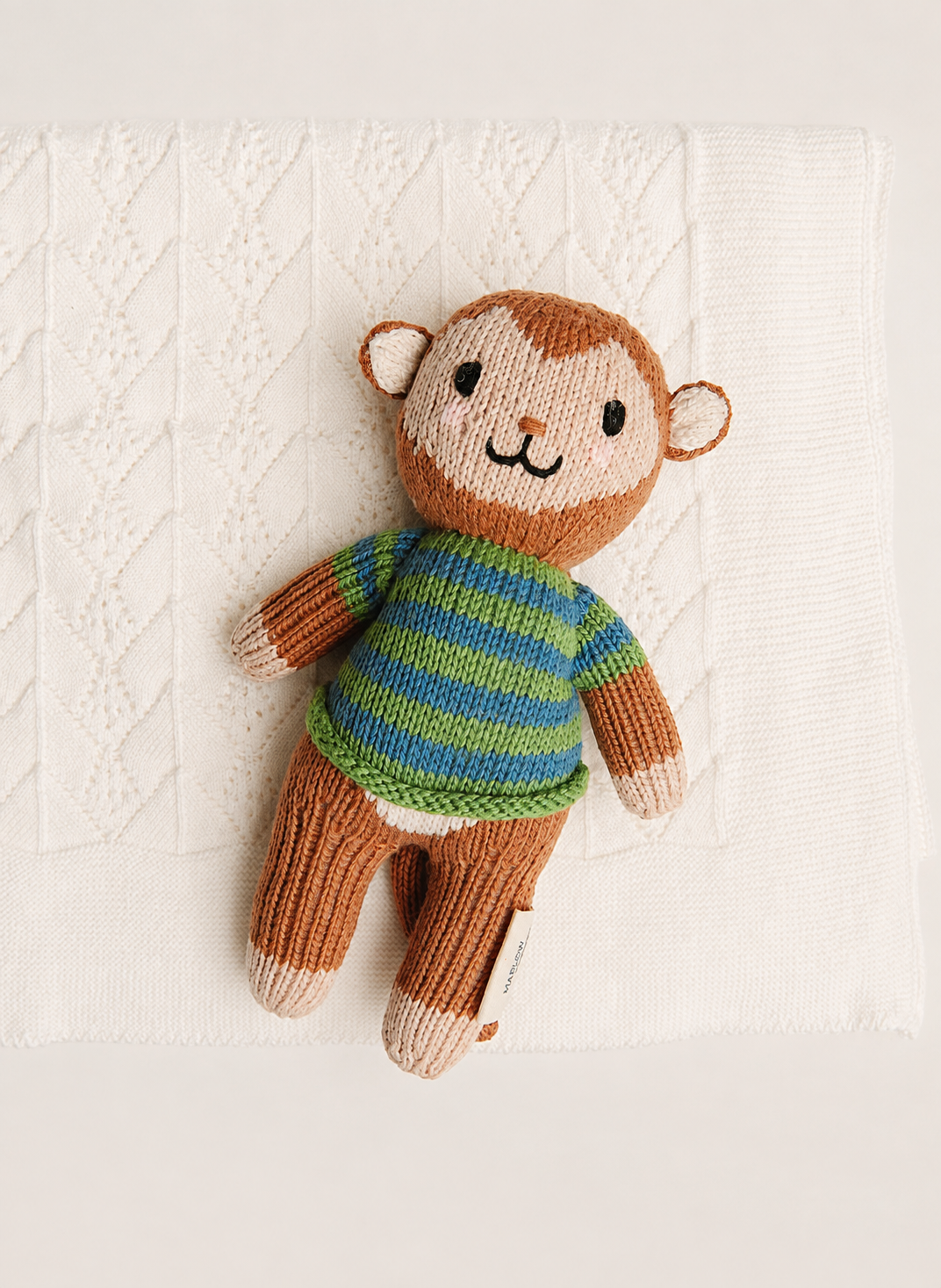 A handmade knitted monkey toy with brown fur-like yarn, a striped green and blue sweater, and cream-colored feet and ears, resting on a textured white knit blanket.