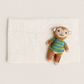 A small knitted monkey toy wearing a green and blue striped sweater, placed on top of a white textured blanket against a warm off-white background.
