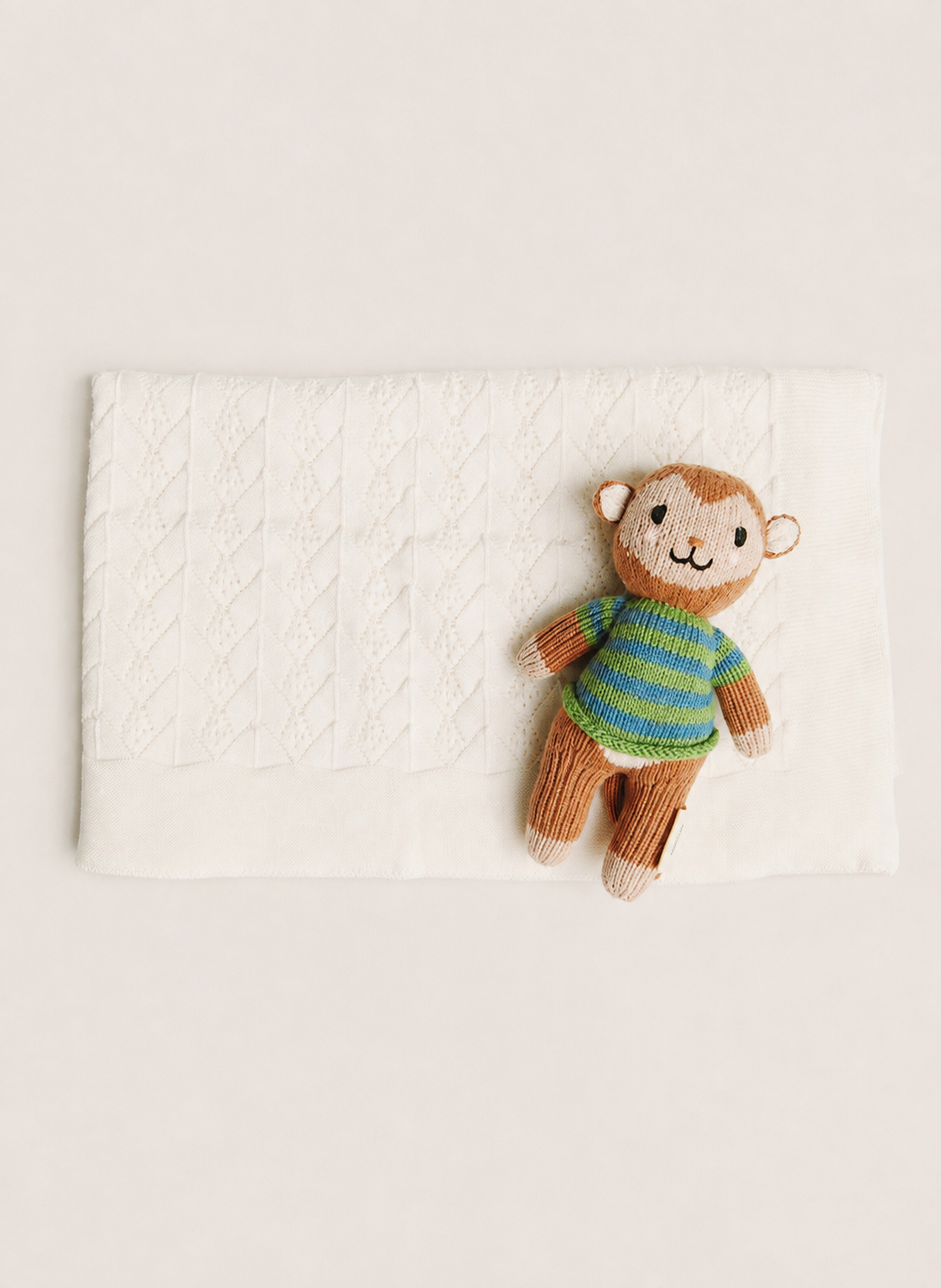 A small knitted monkey toy wearing a green and blue striped sweater, placed on top of a white textured blanket against a warm off-white background.