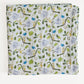 A folded white muslin blanket featuring a colorful repeating pattern of green leaves, blue flowers, and yellow monkeys, displayed against a clean white background.