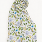 A white muslin baby blanket with a colorful jungle print featuring green leaves, blue butterflies, and yellow monkeys, styled in a spiral fold on a white background.