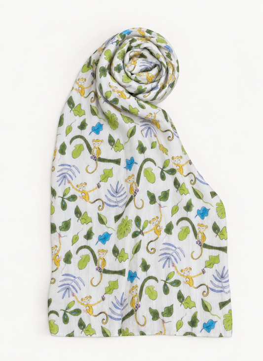 A white muslin baby blanket with a colorful jungle print featuring green leaves, blue butterflies, and yellow monkeys, styled in a spiral fold on a white background.