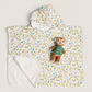 A hooded baby towel or poncho with a white background and a colorful all-over print of dinosaurs and plants. It features a soft white fleece lining on the inside and a small plush teddy bear wearing a green striped sweater placed on the front.