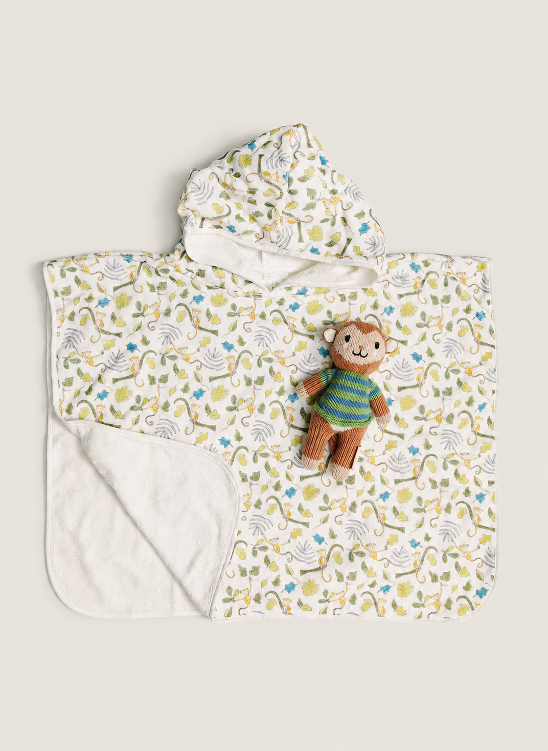 A hooded baby towel or poncho with a white background and a colorful all-over print of dinosaurs and plants. It features a soft white fleece lining on the inside and a small plush teddy bear wearing a green striped sweater placed on the front.