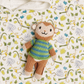 A small knitted monkey toy wearing a striped green and blue sweater, placed on a white quilted blanket with a yellow and green jungle leaf pattern.