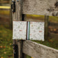 A rectangular fabric pouch with a white background and a multicolor horse and rider pattern, tied to a rustic wooden fence gate outdoors.