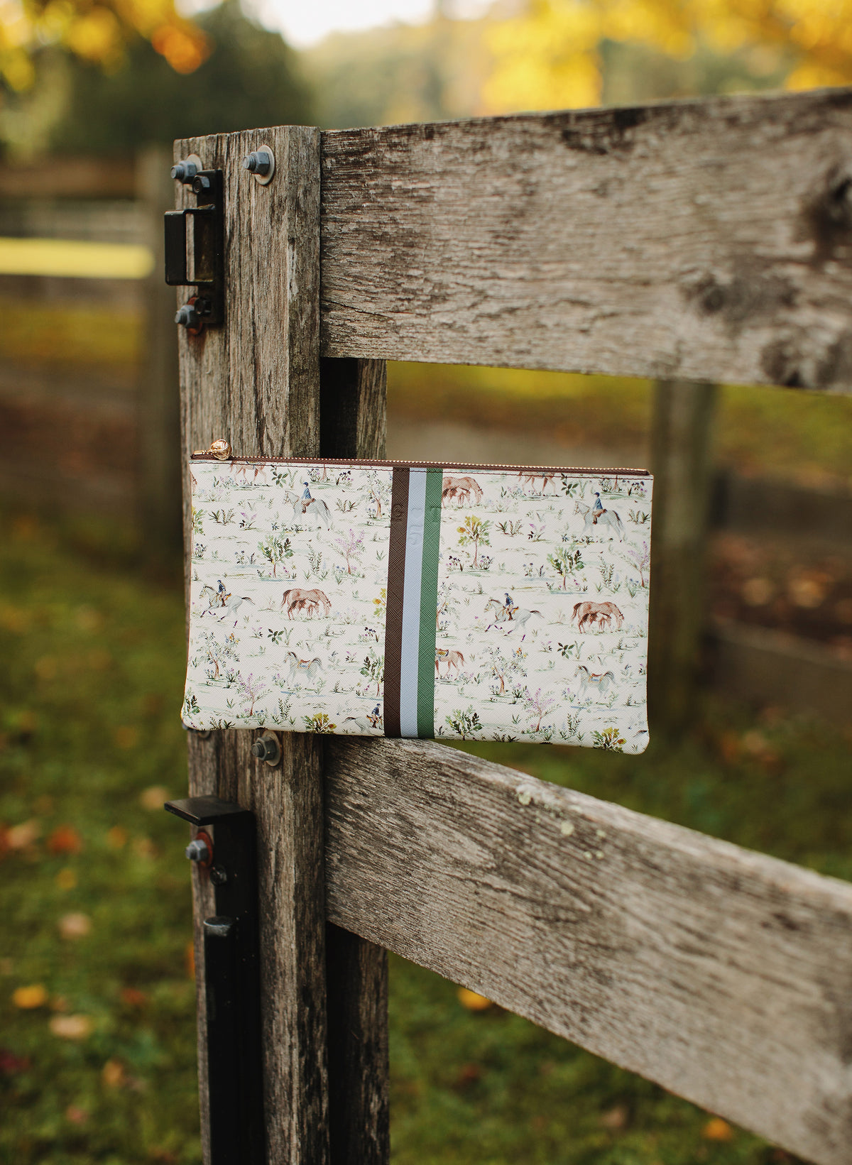 A rectangular fabric pouch with a white background and a multicolor horse and rider pattern, tied to a rustic wooden fence gate outdoors.