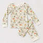A two-piece children's pajama set laid flat on a light beige background. The long-sleeve top and matching pants are cream-colored with an all-over whimsical print of orange-brown cows, green foliage, and small animals.