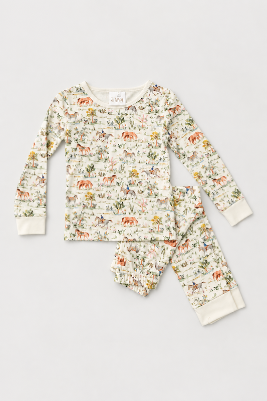 A two-piece children's pajama set laid flat on a light beige background. The long-sleeve top and matching pants are cream-colored with an all-over whimsical print of orange-brown cows, green foliage, and small animals.