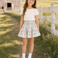 A young girl stands outdoors on a grassy path between a white fence and a rustic barn, wearing a cream short-sleeve top and a flared skirt with a delicate multicolor floral and butterfly print. She is wearing white socks and tan Mary Jane shoes.