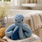 A blue knitted octopus bath toy sits on a cream towel on a wooden stool in a bright bathroom setting with a bathtub, brass faucet, and flowers in a vase.
