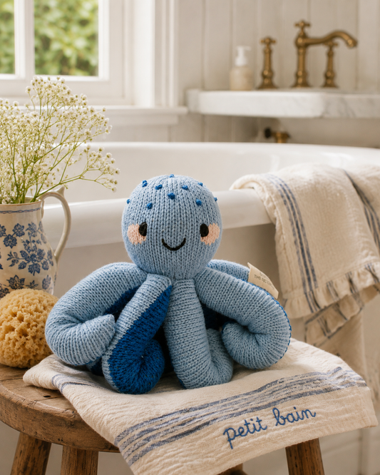A blue knitted octopus bath toy sits on a cream towel on a wooden stool in a bright bathroom setting with a bathtub, brass faucet, and flowers in a vase.