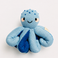 A light blue knitted octopus plush toy with darker blue accents on its tentacles, featuring embroidered eyes, a smile, and small pink cheeks, set against a plain light gray background.