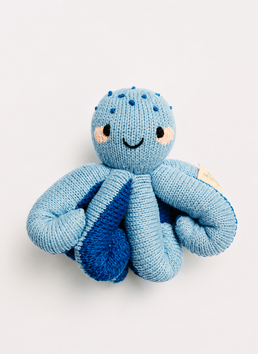 A light blue knitted octopus plush toy with darker blue accents on its tentacles, featuring embroidered eyes, a smile, and small pink cheeks, set against a plain light gray background.