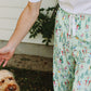 A person wearing light green drawstring pajama pants with a colorful allover ski and tree print, standing outdoors next to a small dog wearing a matching bandana.