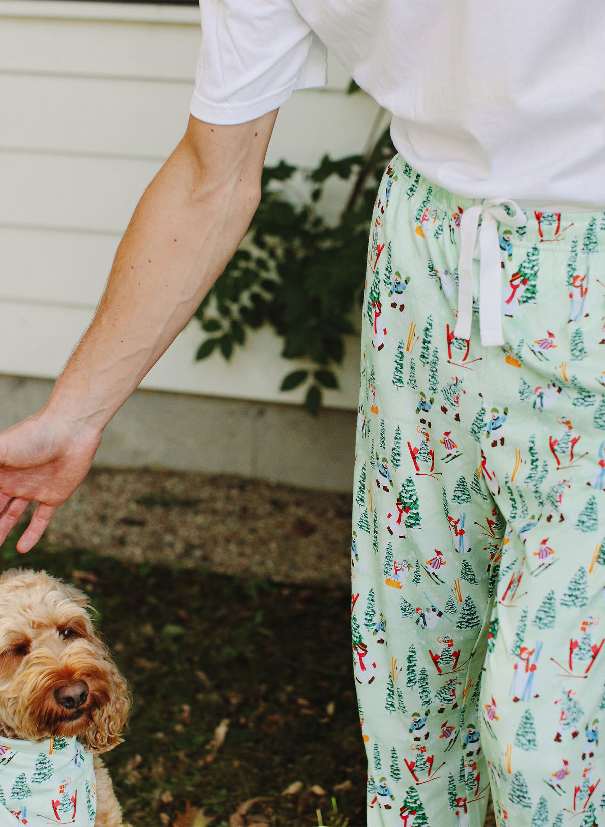 A person wearing light green drawstring pajama pants with a colorful allover ski and tree print, standing outdoors next to a small dog wearing a matching bandana.
