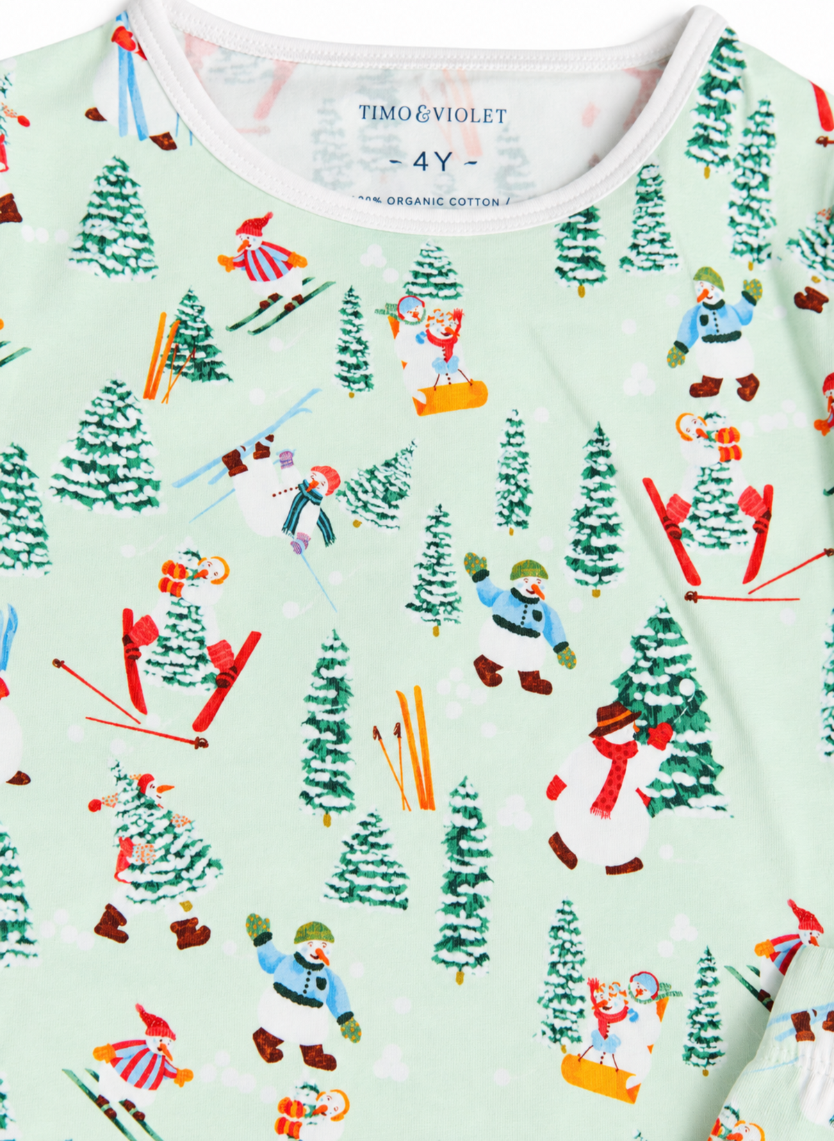A close-up of a children's long-sleeve top by Timo & Violet, featuring a light mint green background with a colorful winter ski print. The pattern includes cartoon snowmen, skiers, and green pine trees.
