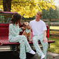 A man and a woman wearing matching light green floral pajama sets sit in the back of a red pickup truck outdoors, with a small dog in a matching pet outfit between them. Trees with yellow autumn leaves and a fence are in the background.