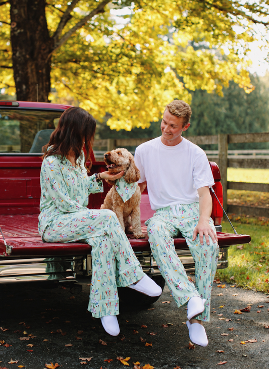 A man and a woman wearing matching light green floral pajama sets sit in the back of a red pickup truck outdoors, with a small dog in a matching pet outfit between them. Trees with yellow autumn leaves and a fence are in the background.