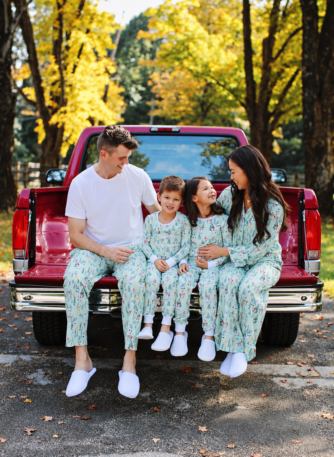 A family of four wearing matching floral print pajamas sits together on the tailgate of a red pickup truck in an outdoor setting with autumn trees in the background.