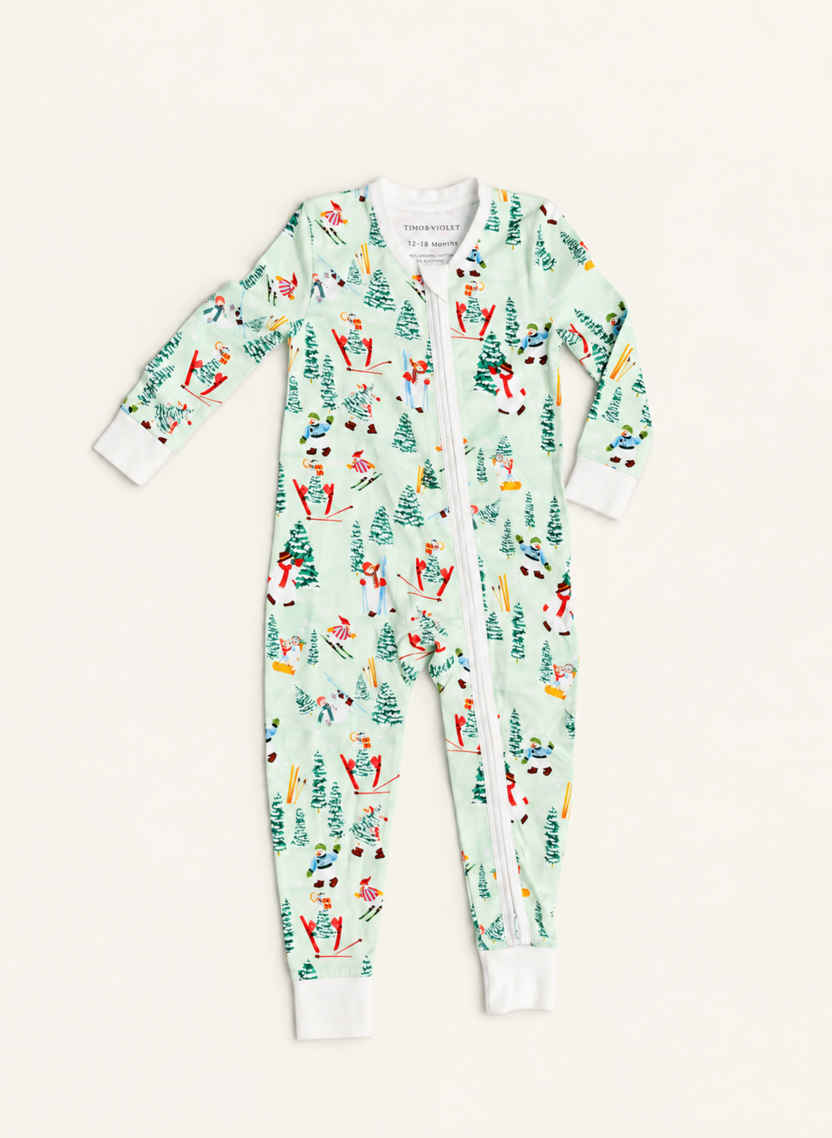 A baby's long-sleeve zip-up footed sleeper with a festive winter print of skiers, snowboarders, and Christmas trees on a pale mint green background, shown flat on a light cream background.