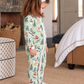 A young girl wearing a light green footed pajama with a colorful winter forest print, standing in a cozy bedroom setting.
