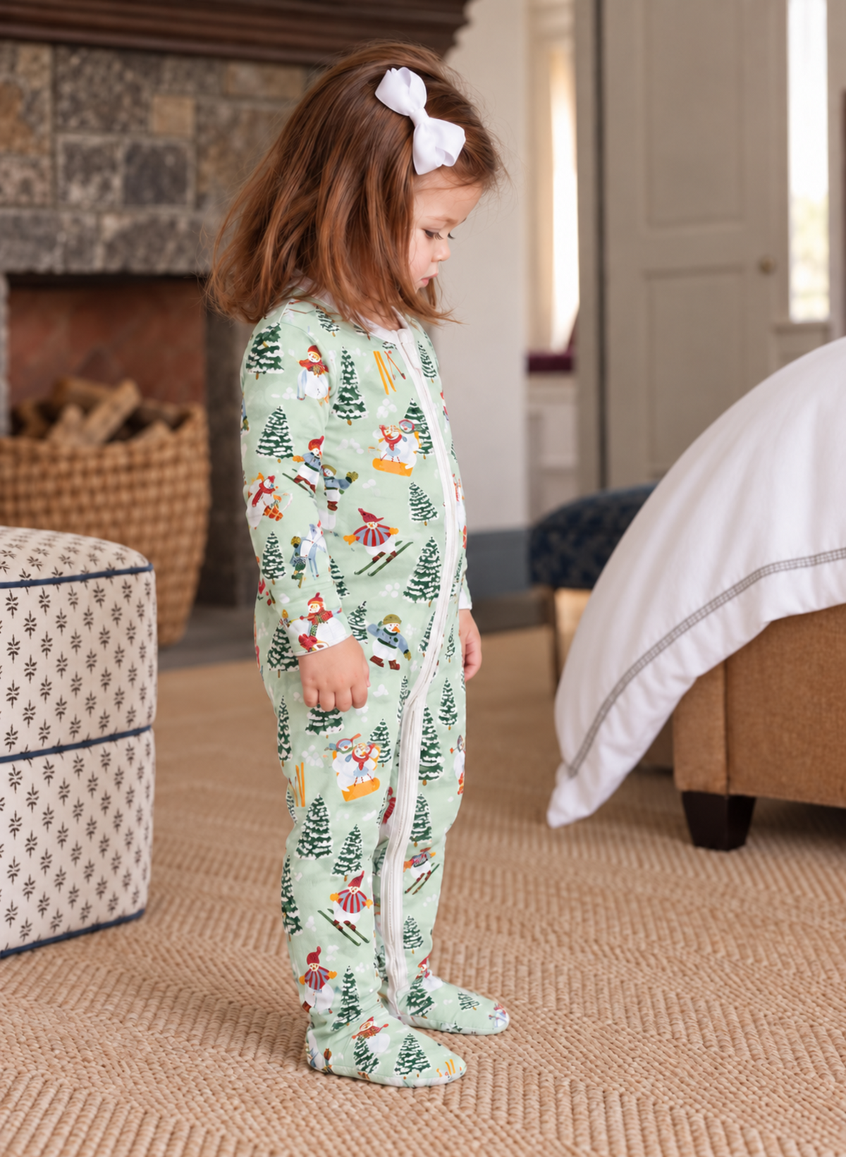 A young girl wearing a light green footed pajama with a colorful winter forest print, standing in a cozy bedroom setting.
