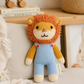 A knitted lion plush toy with an orange yarn mane, yellow arms and legs, and light blue overalls, sitting on a beige cushion in a styled indoor setting.