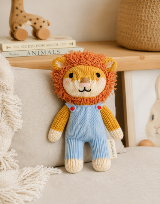 A knitted lion plush toy with an orange yarn mane, yellow arms and legs, and light blue overalls, sitting on a beige cushion in a styled indoor setting.
