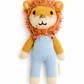 A knitted lion doll with a fluffy orange-brown mane, cream face and paws, yellow arms, and light blue knitted overalls, photographed on a plain white background. A small fabric tag reads TUMI KNOTSART.