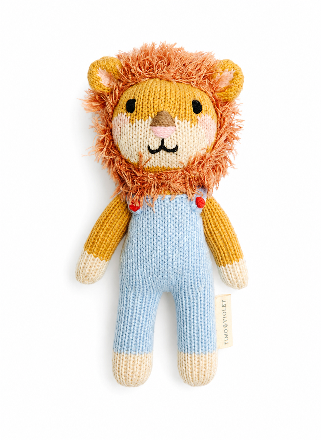 A knitted lion doll with a fluffy orange-brown mane, cream face and paws, yellow arms, and light blue knitted overalls, photographed on a plain white background. A small fabric tag reads TUMI KNOTSART.