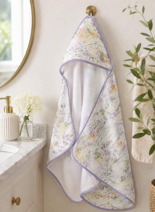A baby hooded towel with a pastel floral print and lavender trim hangs from a gold wall hook in a bathroom setting.