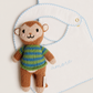 A knitted monkey plush toy wearing a green and blue striped sweater, posed in front of a white bib with light blue scalloped edges and the word 'amore' embroidered on it.