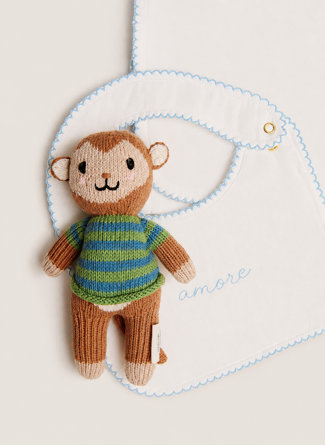 A knitted monkey plush toy wearing a green and blue striped sweater, posed in front of a white bib with light blue scalloped edges and the word 'amore' embroidered on it.