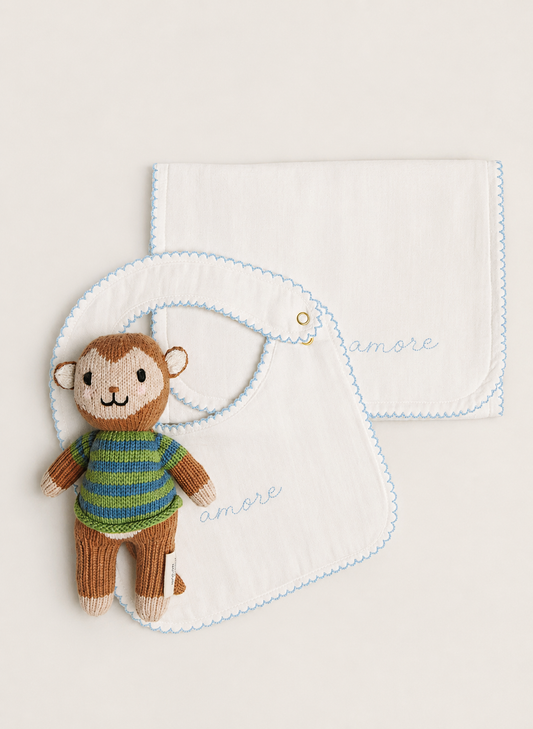 Two white muslin baby bibs with light blue scalloped edges and embroidered 'amore' script, styled with a small knitted monkey doll on a warm off-white background.