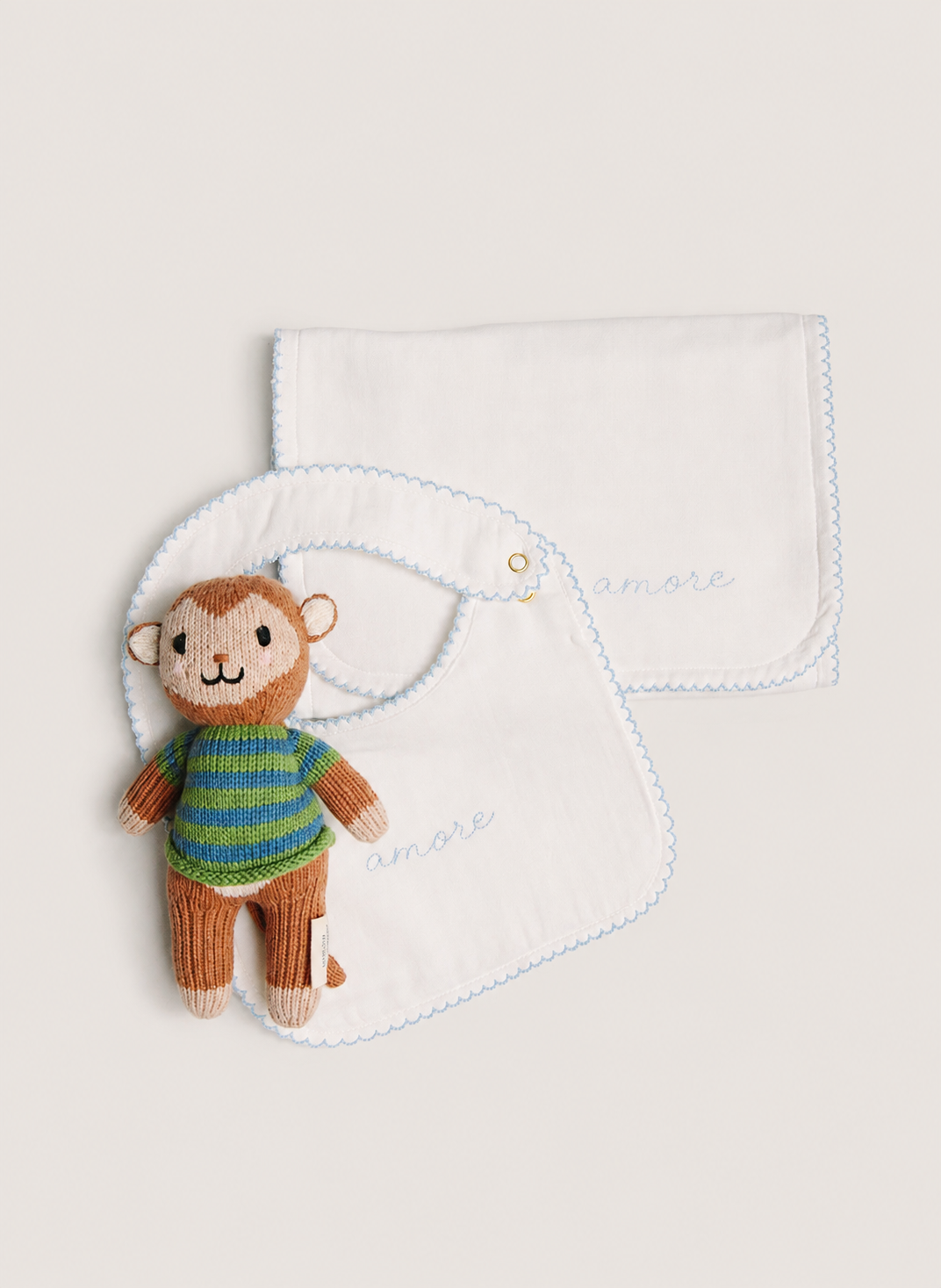 A set of two white baby bibs with light blue scalloped edges, one laid flat and one hanging, featuring the embroidered brand name 'anore'. A small plush monkey toy wearing a green and blue striped shirt is tucked into the hanging bib.