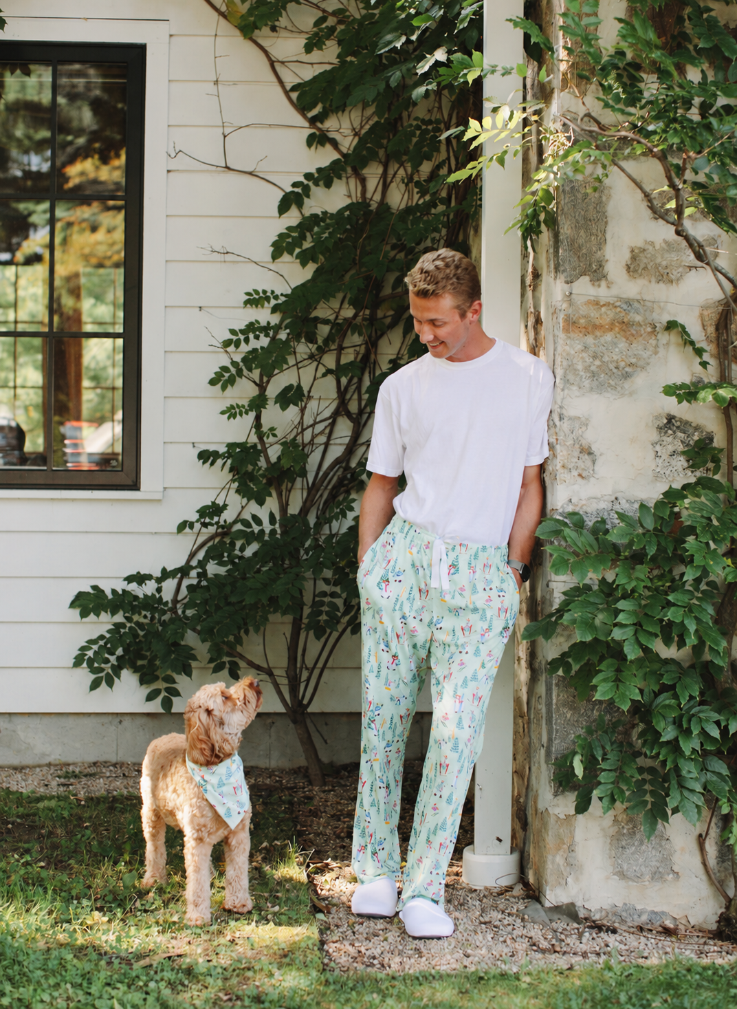 A man wearing a white t-shirt and light green floral print pants stands outdoors next to a stone pillar, with a small dog wearing a matching bandana standing nearby. The background shows a white house exterior with green vines.