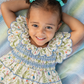 A smiling young girl with curly hair in two buns tied with turquoise bows lies on a striped pastel blanket, wearing a tiered cream dress with a colorful toucan print and blue smocked bodice details.