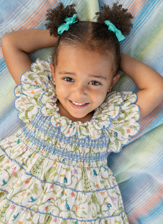 A smiling young girl with curly hair in two buns tied with turquoise bows lies on a striped pastel blanket, wearing a tiered cream dress with a colorful toucan print and blue smocked bodice details.