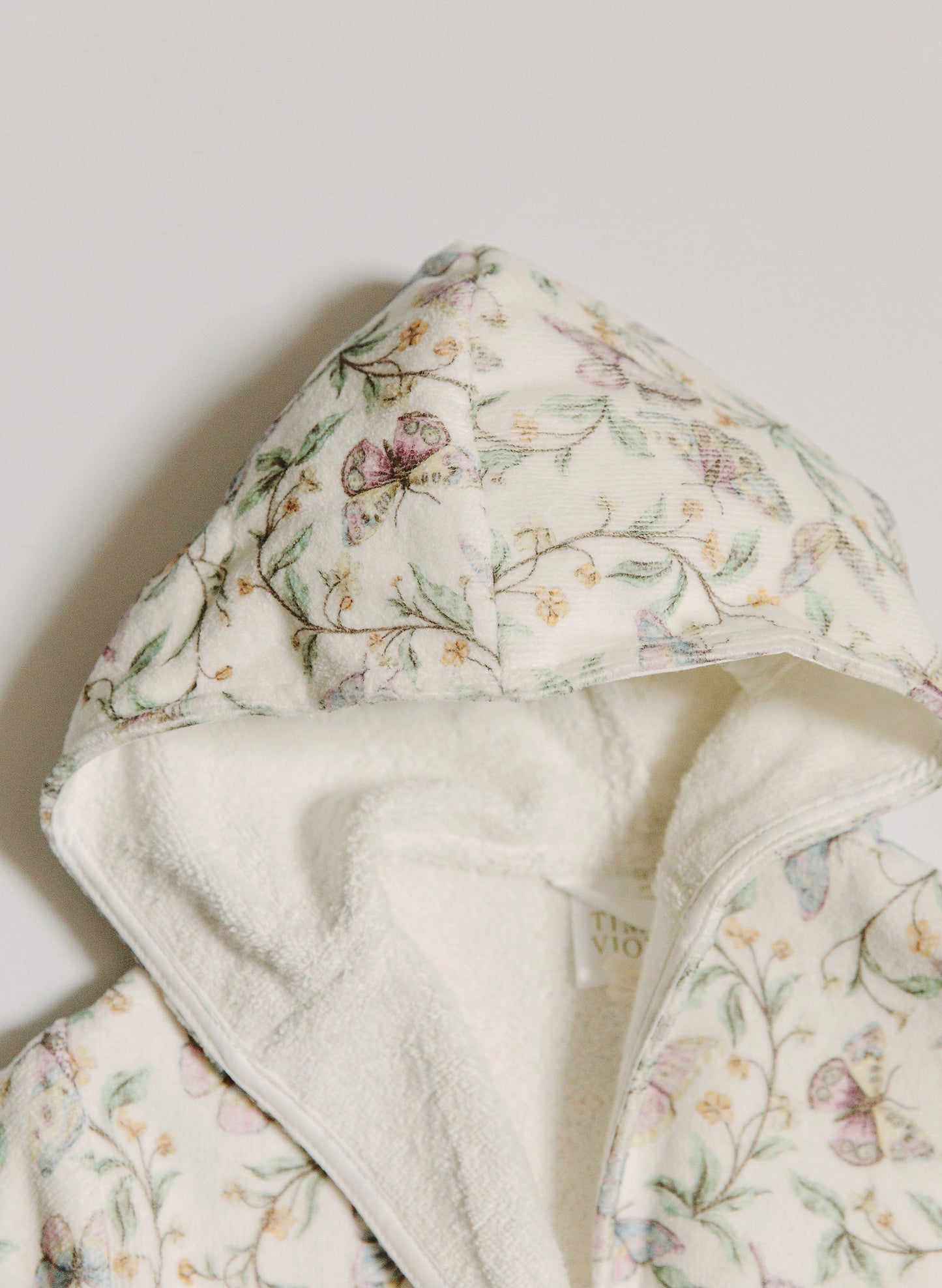 A close-up of a hooded garment, likely a robe or blanket, featuring a cream-colored outer fabric with a delicate pastel floral and butterfly print, and a plush cream sherpa lining.