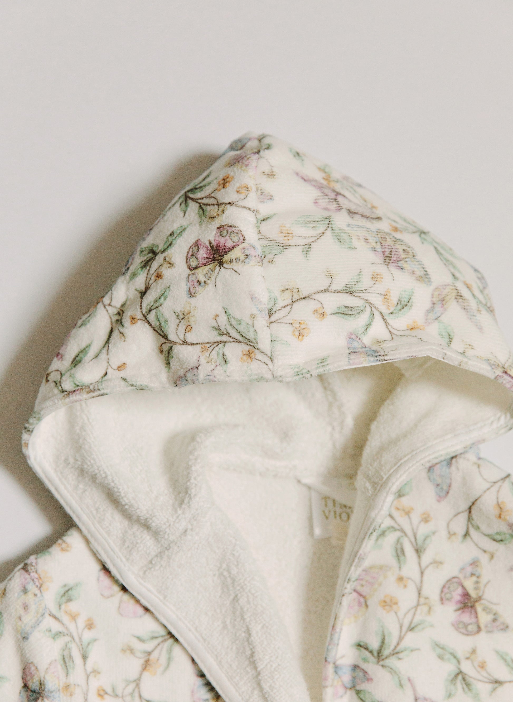 A close-up of a hooded garment, likely a robe or blanket, featuring a cream-colored outer fabric with a delicate pastel floral and butterfly print, and a plush cream sherpa lining.