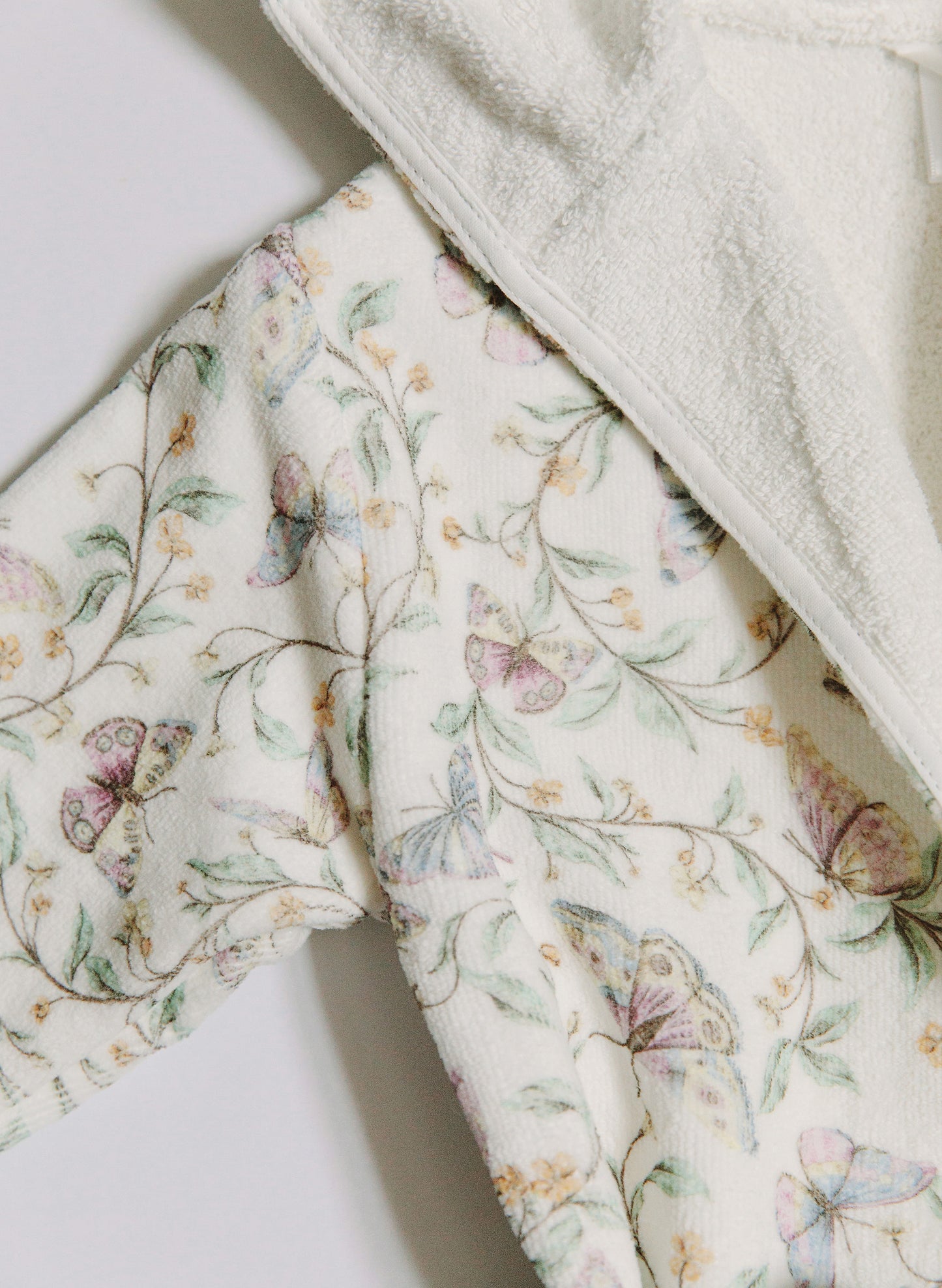 Close-up of a soft fleece or plush robe with a white border and a delicate pastel butterfly and floral print on an off-white background.