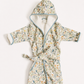 A children's hooded bathrobe with a tie belt, featuring a delicate multicolor floral and botanical print on a light cream base, with light blue piping and a soft cream lining.