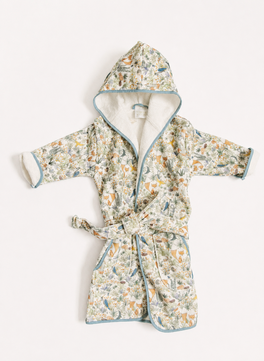 A children's hooded bathrobe with a tie belt, featuring a delicate multicolor floral and botanical print on a light cream base, with light blue piping and a soft cream lining.