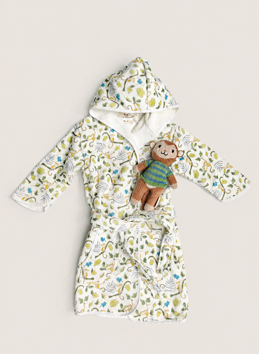 A baby hooded bathrobe with a colorful monkey and leaf pattern on a cream background, styled with a small plush monkey toy, laid flat on a light beige background.